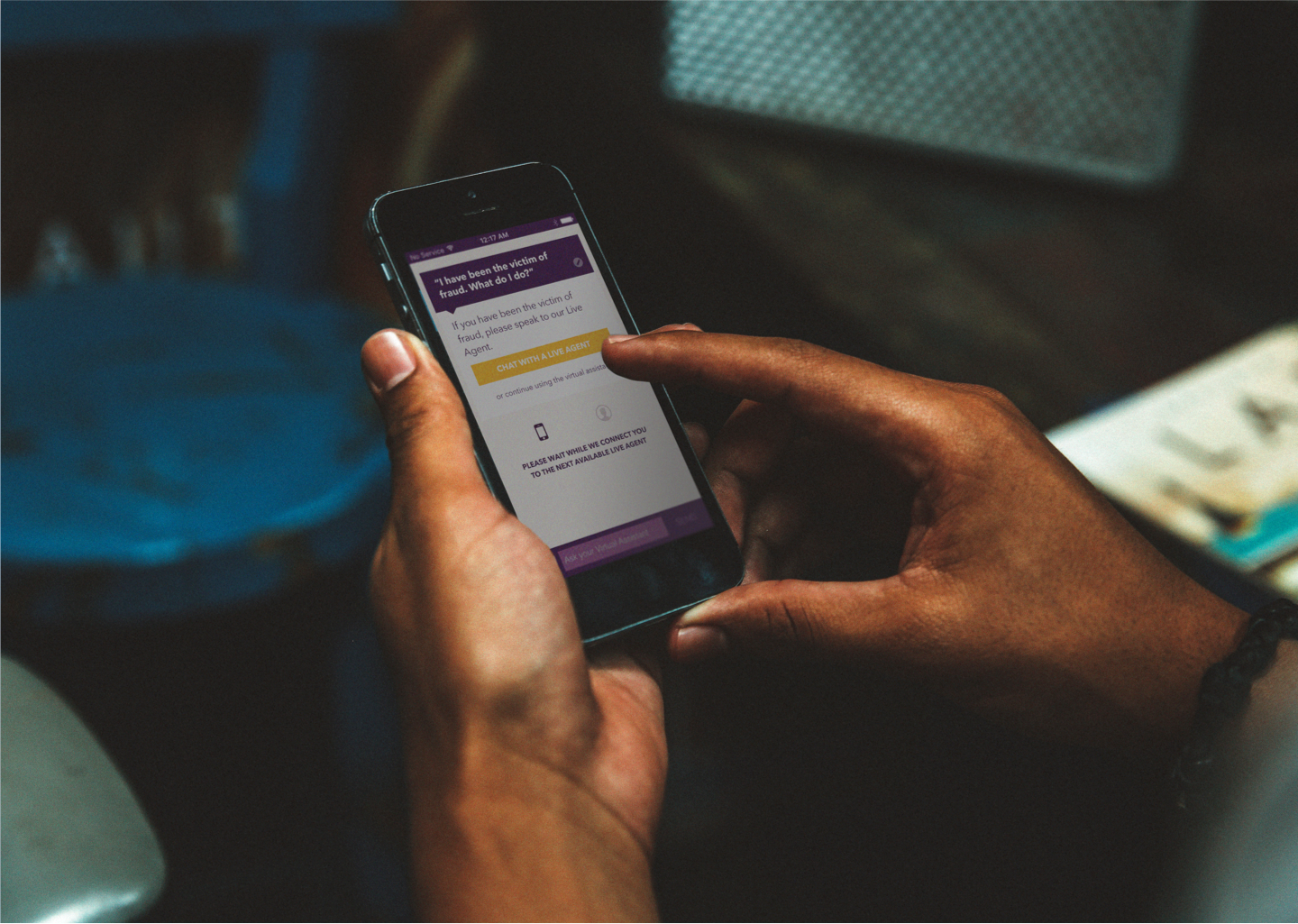 Close-up photo of hands interacting with a mobile phone displaying the Kasisto App virtual assistant interface. The screen is displaying a conversation about fraud and the user is clicking the chat with a live agent button.